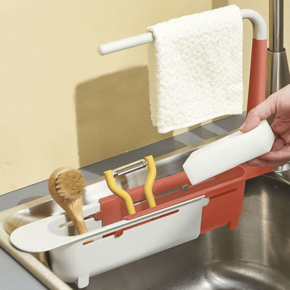 Red and white telescopic sink rack holding a cloth brush peeler and sponge by a stainless steel sink